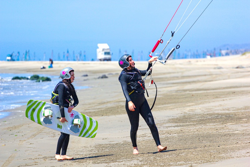 A kitesurf student walking with the kite while another student carries her board upwind in Peru